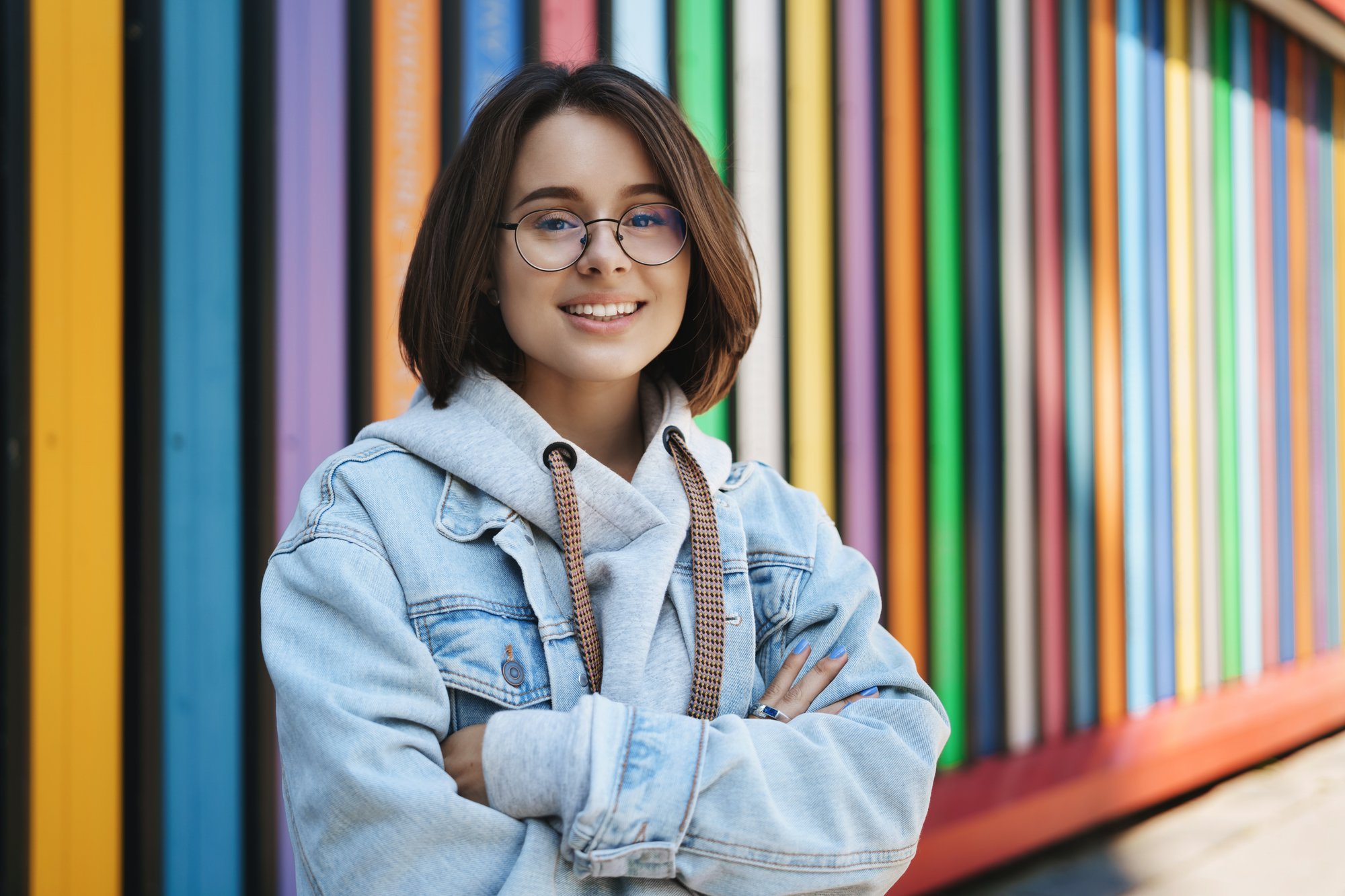 cheerful-young-queer-girl-glasses-denim-jacket-standing-near-rainbow-wall-cross-hands-chest-smiling-camera-happy-lifestyle-urban-life-generation-concept