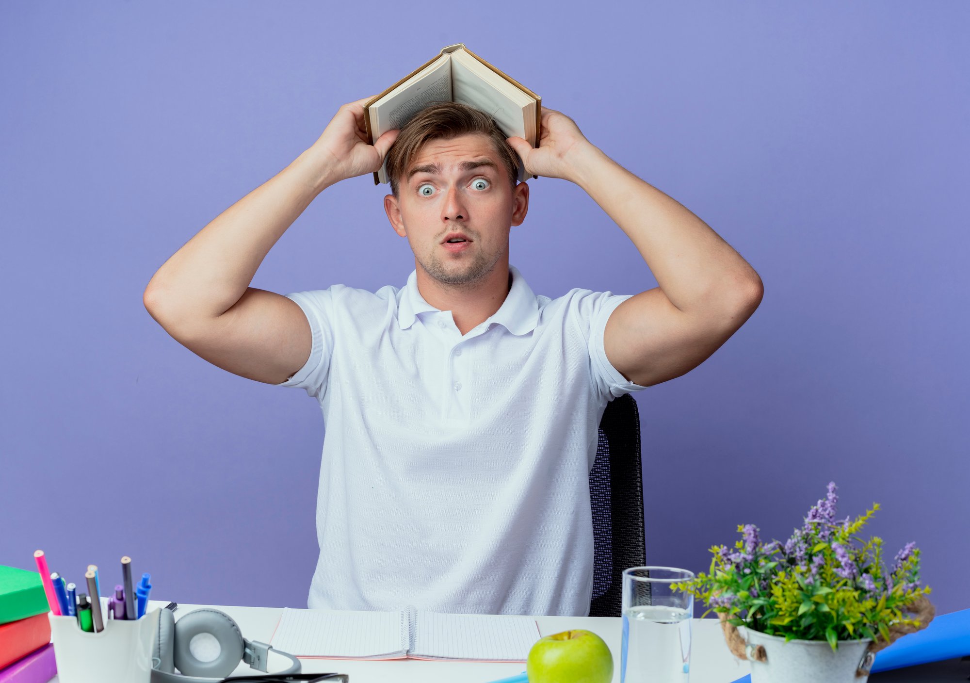 surprised-young-handsome-male-student-sitting-desk-with-school-tools-covered-head-with-book-isolated-blue